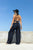 Person wearing a black outfit with wide-leg pants and a hat, standing against a clear blue sky.